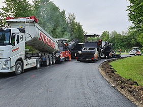 Foto von einem STRABAG LKW beim Abladen, daneben eine Asphaltbaumaschine
