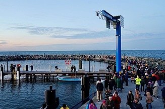 Eine Hand voll Menschen befindet sich auf einer Seebrücke. Die Seebrücke ist abgerundet und ein blaue Säule steht in der Mitte, sie ebnet den Weg zu einem Steg, der in die Mitte der Seebrücke führt.