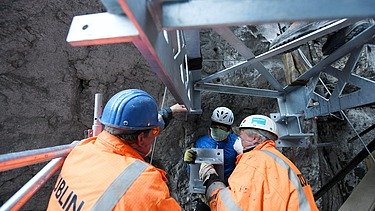Foto von drei Personen in Schutzuniform und Mund-Nase Maske die an einem Stahlträger arbeiten.     