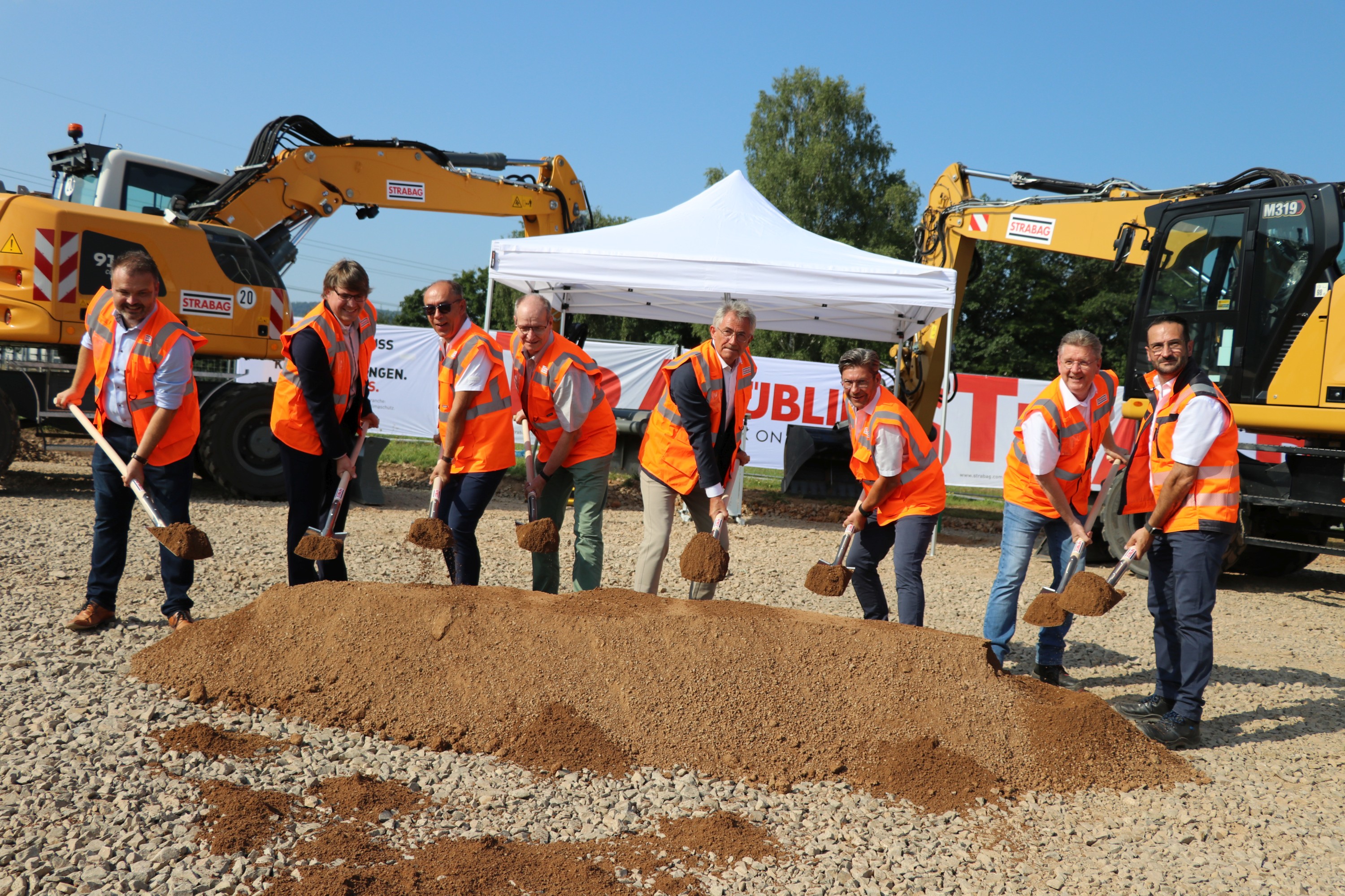 Foto von mehreren Personen bei sonnigem Wetter auf einer Baustelle, die jeweils einen Spaten in den Händen halten und einen symbolischen ‚Spatenstich‘ ausführen