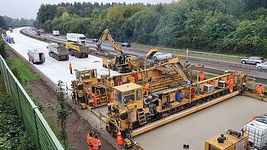 The picture shows a large concrete road construction machine in operation