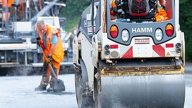 Foto einer Straßenwalze, dahinter steht ein Arbeiter mit einer Schaufel