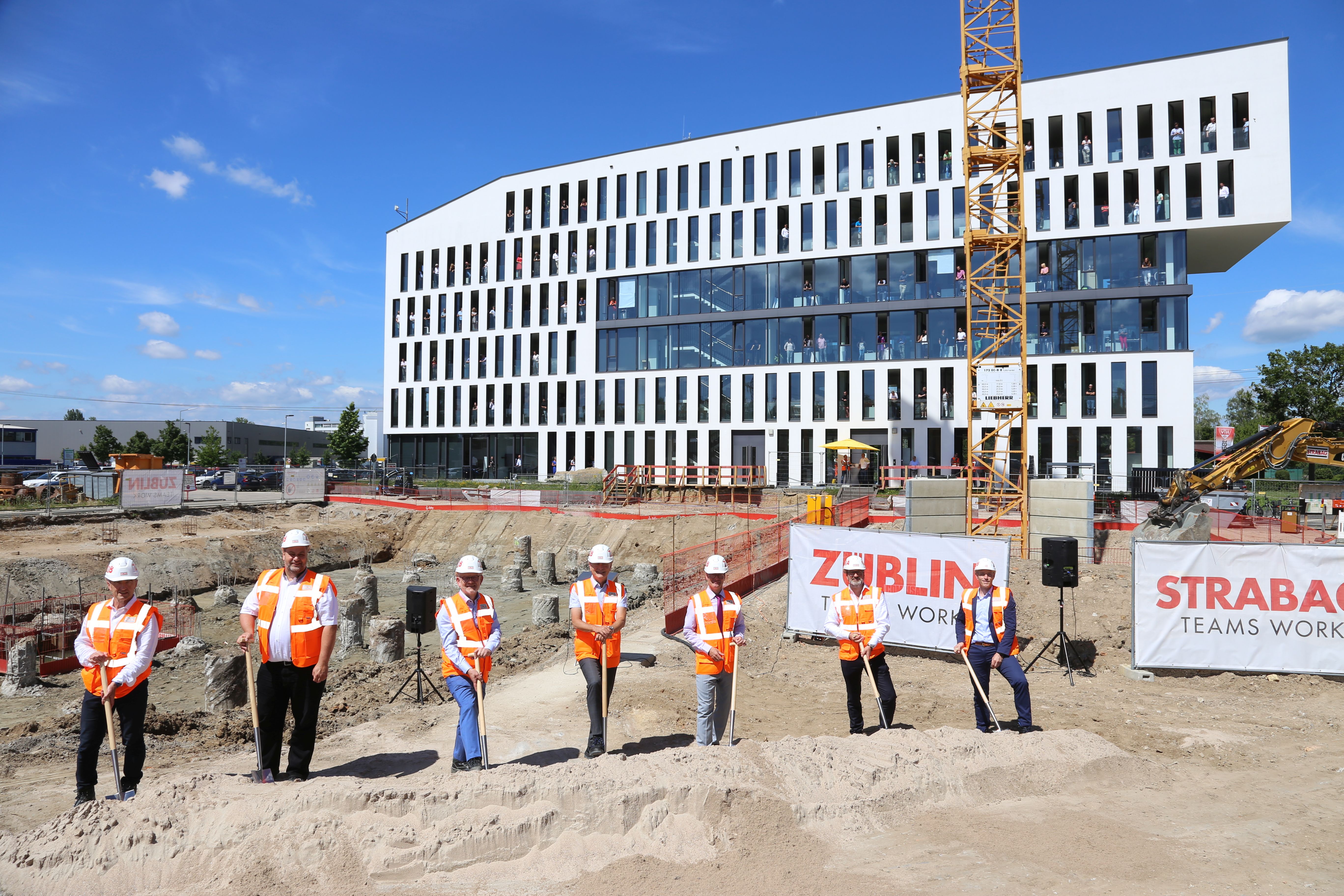 Foto von mehreren Männern in Warnweste auf einer Baustelle. Sie halten einen Spart in der Hand, der in den Boden gesteckt ist. Die Baustelle befindet sich vor einem Bürogebäude. 