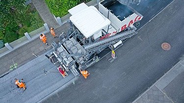 Luftaufnahme einer Asphaltbaumaschine, einige Straßenbauarbeiter stehen an den Seiten