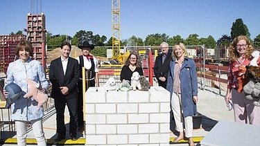 Foto von mehren Personen auf einer Baustelle, sie sind schick gekleidet und haben Kuscheltiere in der Hand. Sie stehen um eine kleine Mauer auf denen ebenfalls Kuscheltiere liegen.  