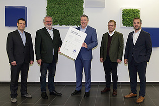 Foto von fünf stehenden Personen, die Person in der Mitte hält eine weiße Tafel in der Hand