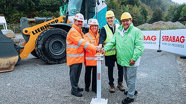 Photo of four people wearing construction helmets and protective jackets standing in front of a wheeled excavator