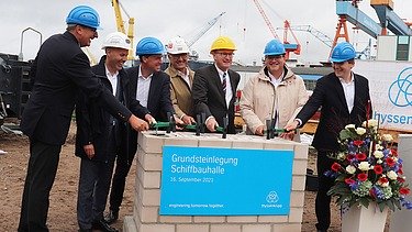 Foto von der Grundsteinlegung. Mehrer Männer stehen um einen Haufen aus Steinen.  