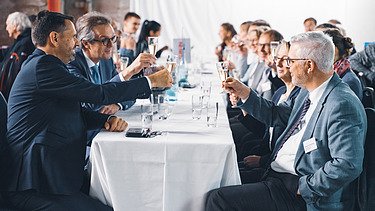 Foto von mehreren Personen bei der Jubiläumsfeier am Standort Sande. Sie sitzen an einem Tisch und stoßen an. 