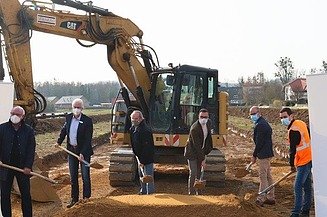 Foto von mehreren Personen beim symbolischen Spatenstich mit Schaufeln in der Hand, dahinter ein großer, gelber Bagger