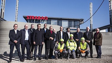 Photo of several people standing in front of a concrete wall, an office building in the background