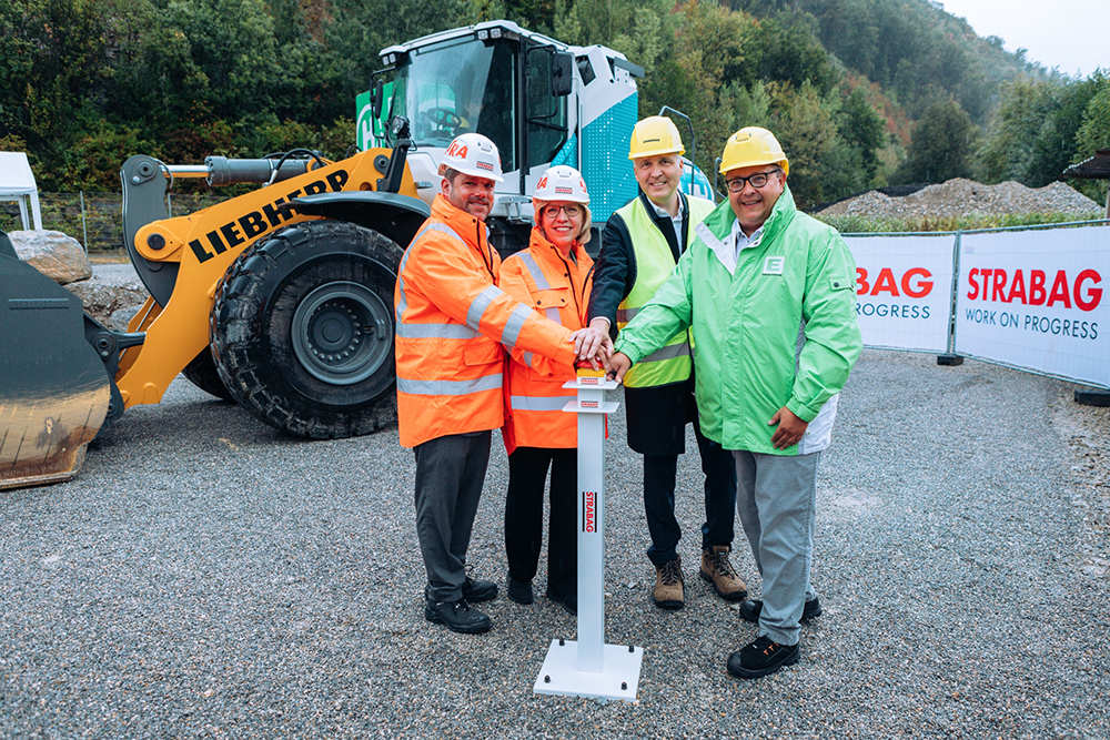 Photo of four people wearing construction helmets and protective jackets standing in front of a wheeled excavator