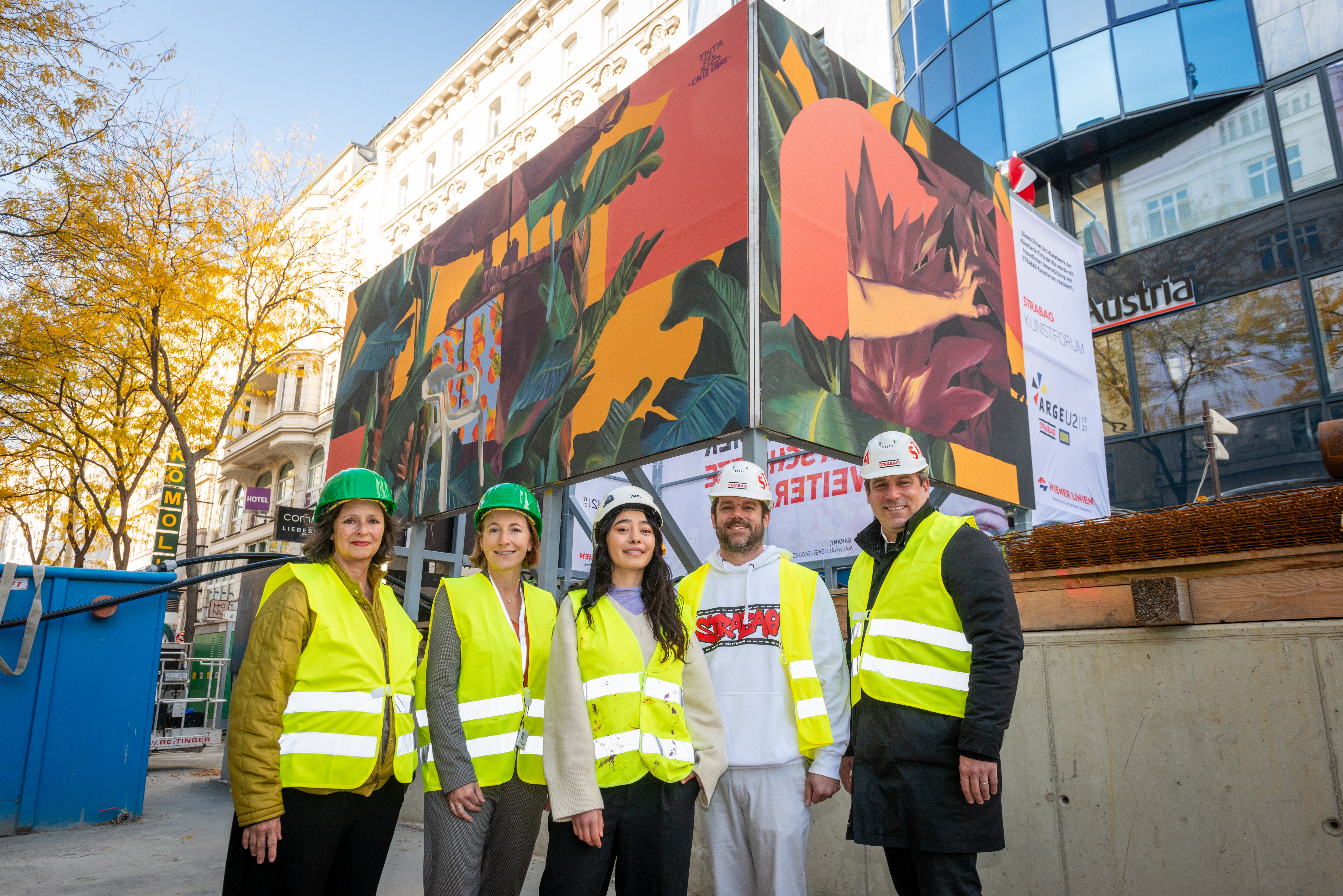 Foto von fünf Personen mit gelben Schutzwesten und Bauhelmen vor einer Baustelle, die mit einem Kunsttransparent umhüllt ist