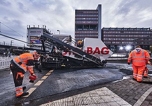 Das Bild zeigt Asphaltierarbeiten mit zwei Arbeitern in Schutzanzügen und einer großen Asphalbaumaschine, im Hintergrund ein Bürogebäude