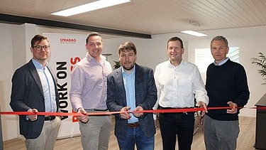 Foto von fünf Männern die ein rotes Band in einem Büroroaum durchschneiden