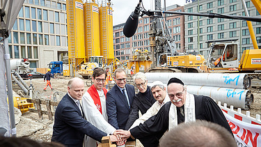 Foto von sechs Männern, darunter drei Priester, auf einer Baustelle, die gemeinsam einen Buzzer drücken. 