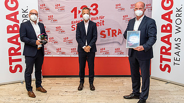 Foto von drei Personen, stehend mit Covid-Masken, die rechte Person hält eine Urkunde in der Hand, im Hintergrund Roll-Ups der STRABAG