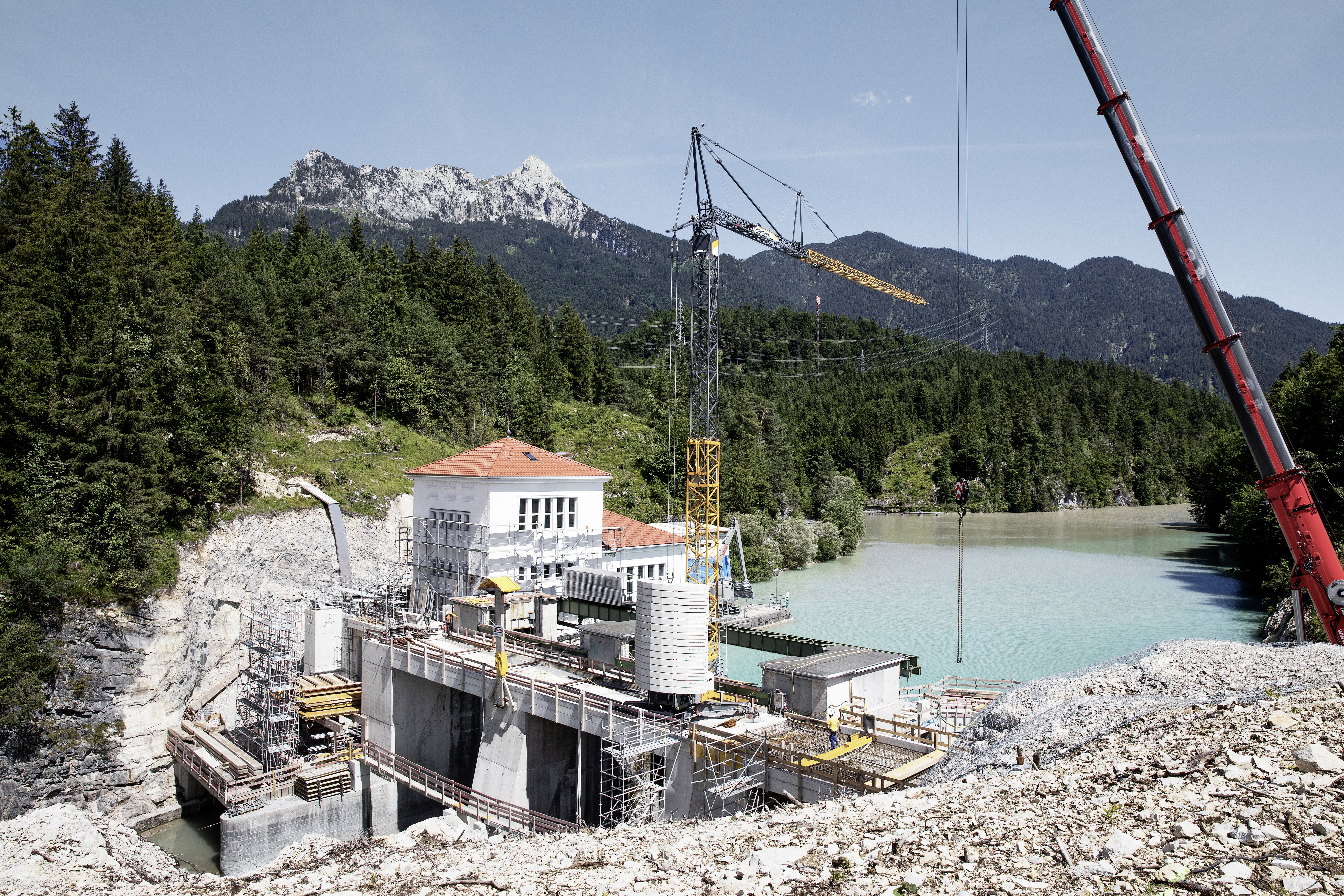 Das Foto zeigt die Baustelle eines Damms in Tirol vor einem See in einem Waldgebiet.