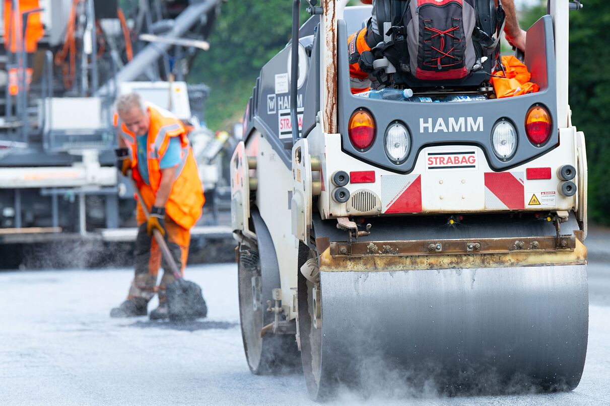 Foto einer Straßenwalze, dahinter steht ein Arbeiter mit einer Schaufel