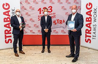 Foto von drei Personen, stehend mit Covid-Masken, die rechte Person hält eine Urkunde in der Hand, im Hintergrund Roll-Ups der STRABAG