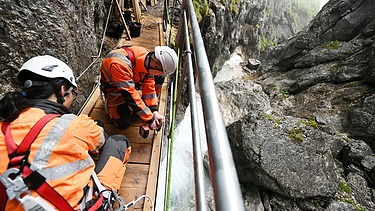 Foto von zwei Personen in Schutzuniform die in einer Schlucht an einer kleinen Brücke schrauben.    