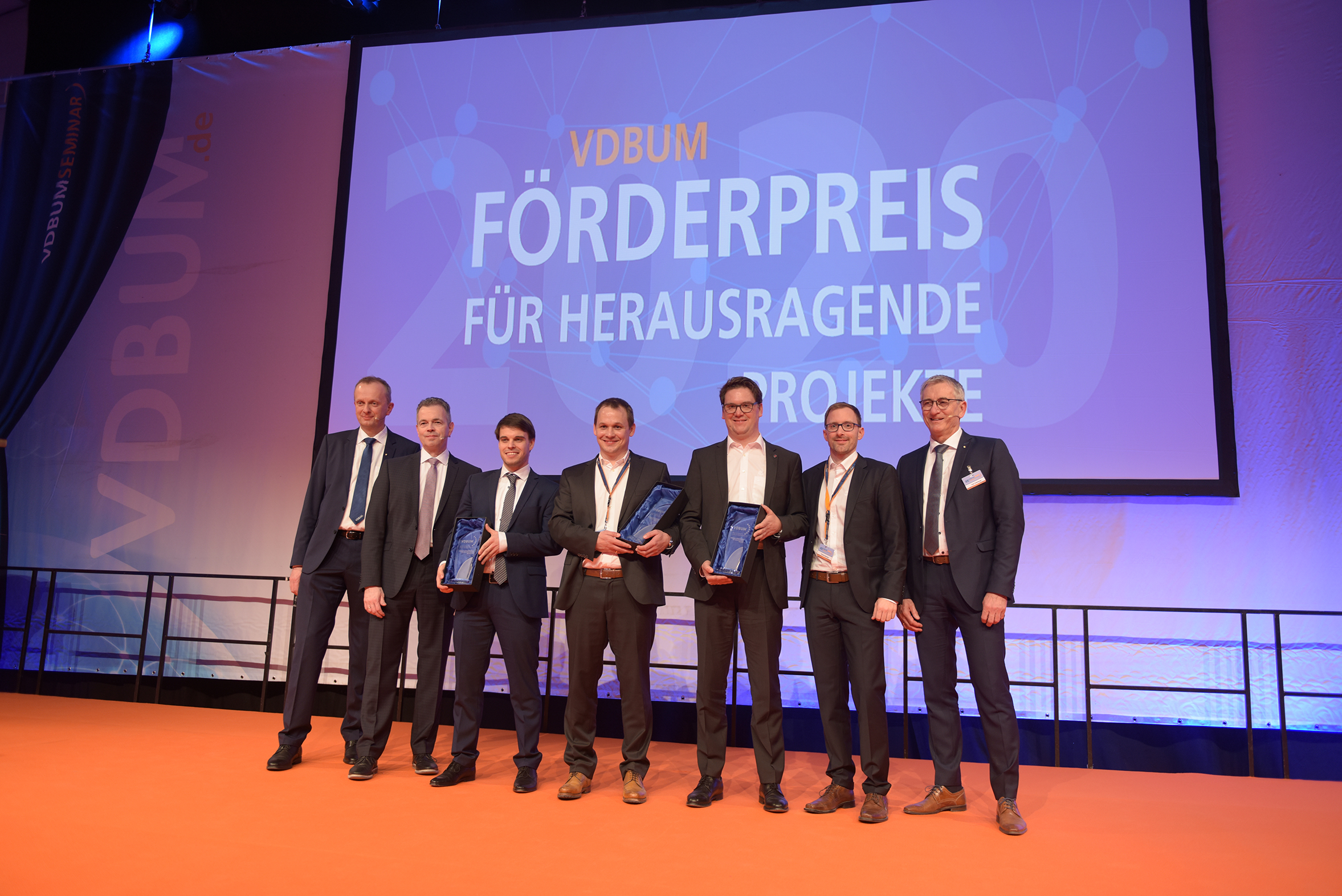 Foto von mehreren Männern in Anzug auf einem Bühnen. Im Hintergrund ein Banner vom VDBUM: „Förderpreis für Herausragende Projekte“ zu sehen.            