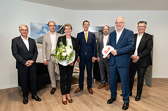Foto von mehren Personen die schick gekleidet sind. Sie schauen freundlich. Vorne steht eine Frau mit Blumen in der Hand. 