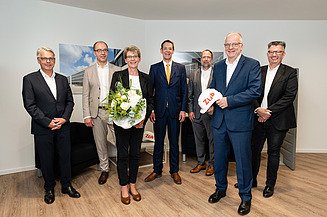 Foto von mehren Personen die schick gekleidet sind. Sie schauen freundlich. Vorne steht eine Frau mit Blumen in der Hand. 
