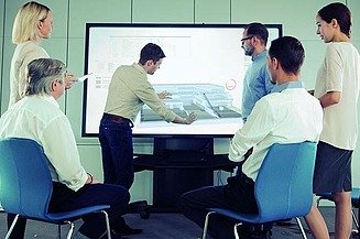 Foto von mehren Personen, die vor einem Fernseher stehen. Es ist eine Präsentation von dem Modele eines Hauses zusehen. Eine Personen verdeutlicht etwas bei der Präsentation.         