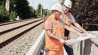 Foto von einem Bahngleis, rechts stehen zwei Personen in Schutzkleidung, die einen Plan in der Hand halten