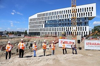 Foto von mehreren Männern in Warnweste auf einer Baustelle. Sie halten einen Spart in der Hand, der in den Boden gesteckt ist. Die Baustelle befindet sich vor einem Bürogebäude. 