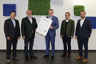 Foto von fünf stehenden Personen, die Person in der Mitte hält eine weiße Tafel in der Hand