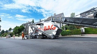 Foto einer Asphaltbaumaschine mit der Aufschrift STRABAG, sie steht am Straßenrand, der Himmel ist bewölkt