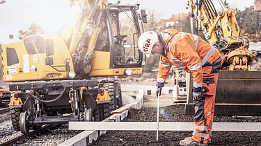 Foto von einem Gleisbauarbeiter in Schutzkleidung, dahinter eine gelbe Gleisbaumaschine