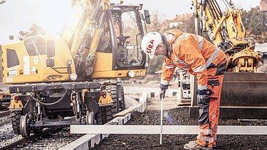 Photo of a track construction worker in protective clothing, behind him a yellow track construction machine
