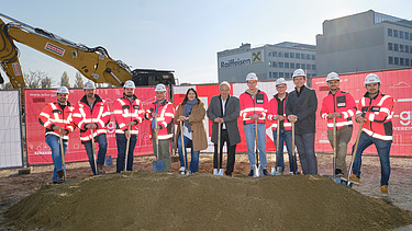 Foto von elf Personen, die einen Spatenstich durchführen, dahinter sieht man einen Bagger und zwei Häuser