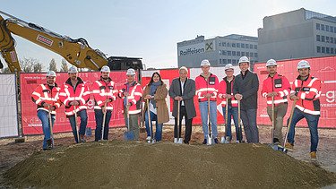 Photo of eleven people breaking ground, behind them you can see an excavator and two houses