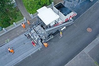 Luftaufnahme einer Asphaltbaumaschine, einige Straßenbauarbeiter stehen an den Seiten