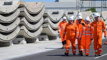 Photo of several people wearing hard hats and orange protective clothing walking next to precast concrete elements