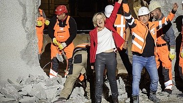 Foto von Personen in Schutzausrüstung und eine schick gekleidete Frau vor einem Tunnel. Die Frau und ein Mann reißen die Arme hoch und freuen sich.  