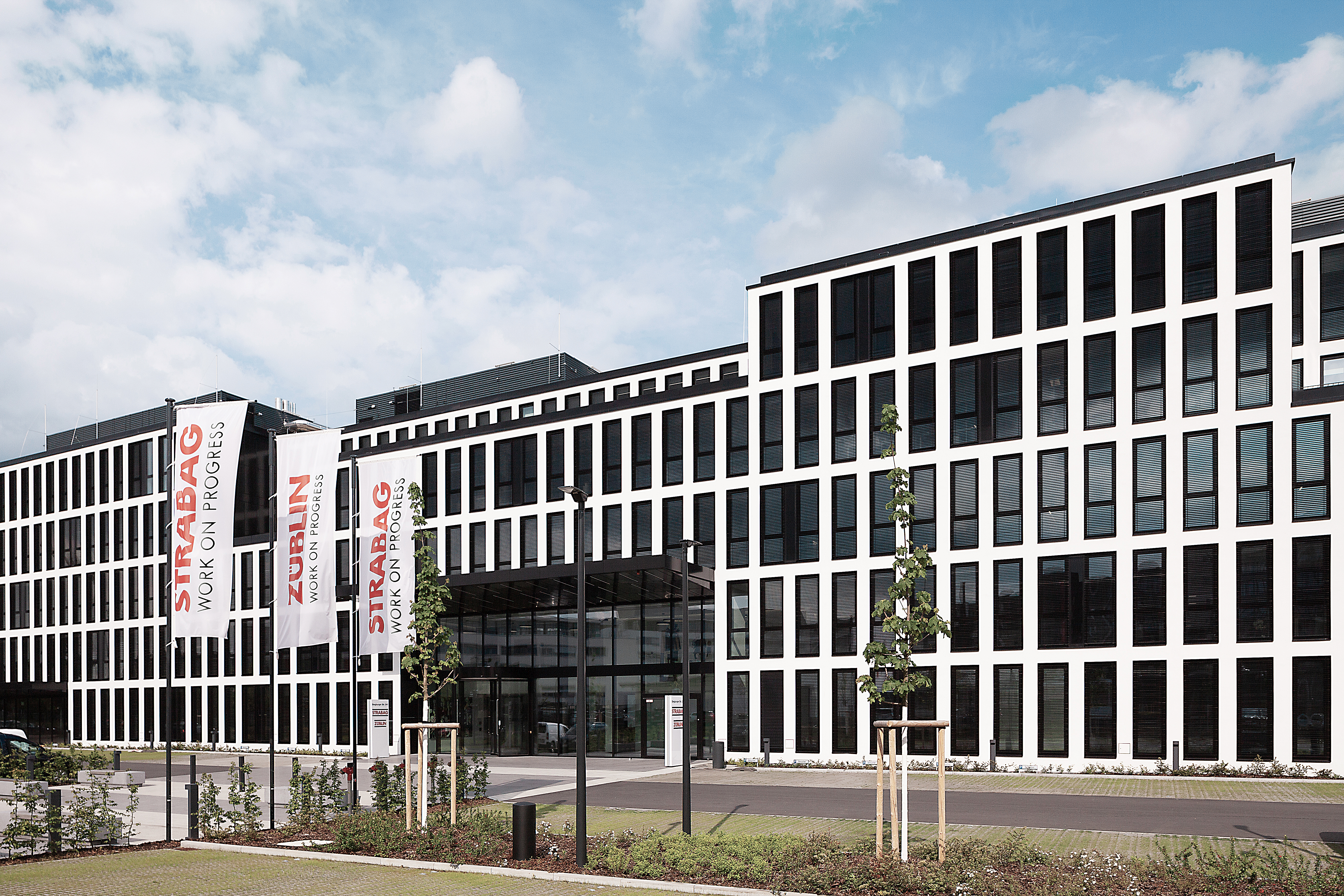 The photo shows the Office at the Cologne site with STRABAG and ZÜBLIN flags in front of the building.