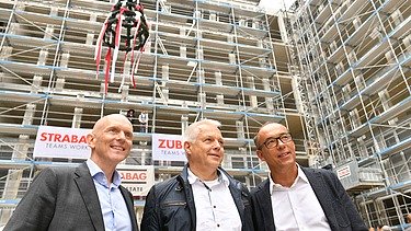 Foto von drei lächelnden Personen auf einer Rohbaustelle. 