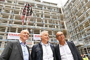Foto von drei lächelnden Personen auf einer Rohbaustelle. 