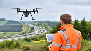 Foto einer Person in Schutzjacke mit einem Tablet in der Hand bedient er eine fliegende Drohne