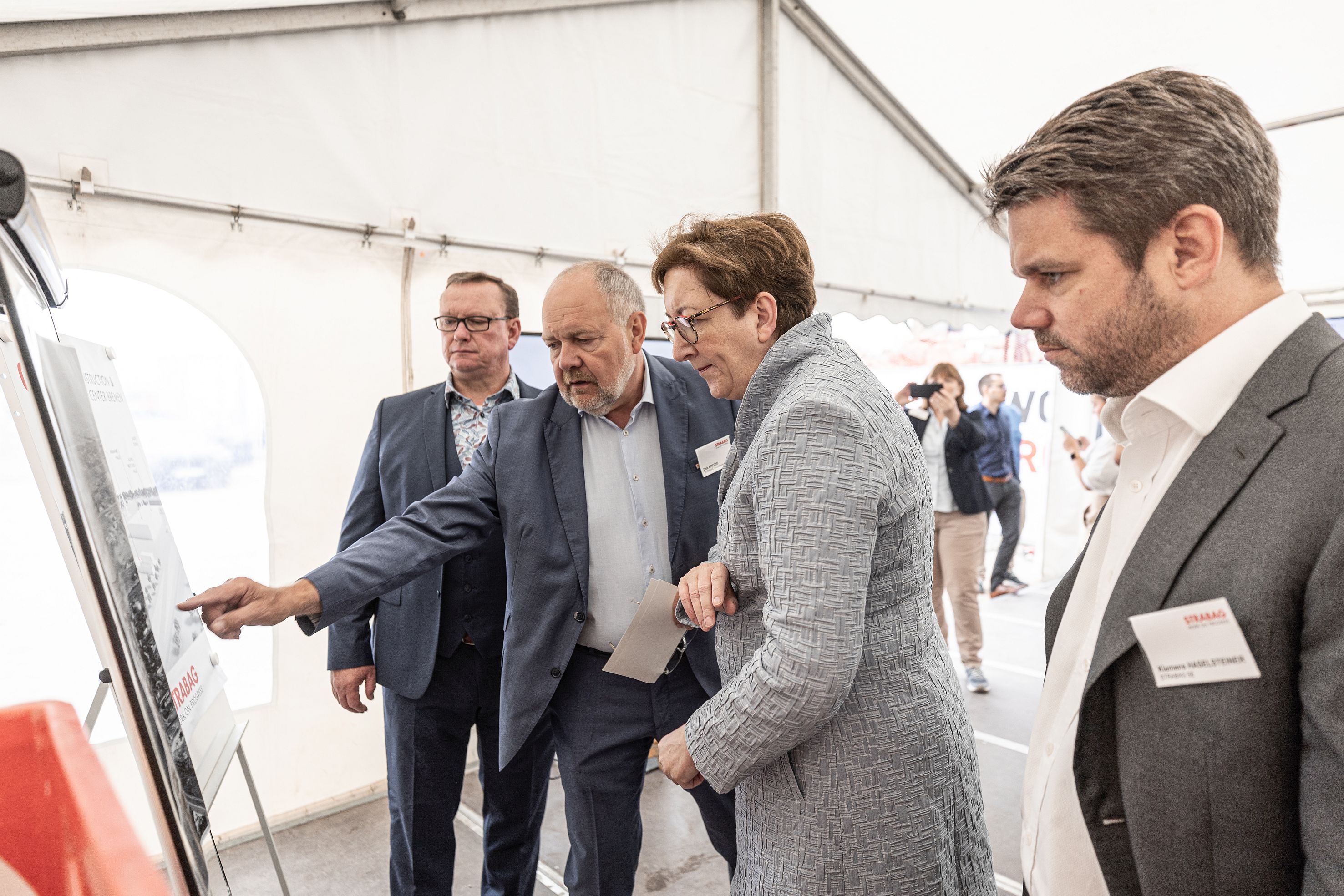 Foto von vier Personen, die eine Bautafel betrachten, die zweite Person von links zeigt darauf