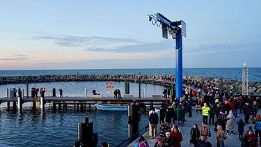 Eine Hand voll Menschen befindet sich auf einer Seebrücke. Die Seebrücke ist abgerundet und ein blaue Säule steht in der Mitte, sie ebnet den Weg zu einem Steg, der in die Mitte der Seebrücke führt.