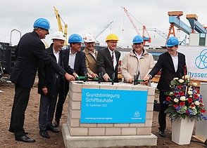 Foto von der Grundsteinlegung. Mehrer Männer stehen um einen Haufen aus Steinen.  