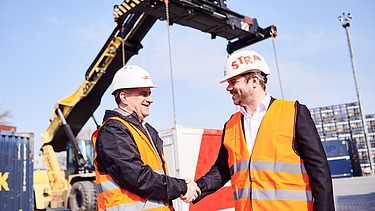 Photo of two people shaking hands and wearing protective clothing, behind which you can see an excavator with its gripper arm extended