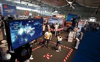 Foto von einem Großbildschirm in einer Messehalle, davor stehen zwei Personen