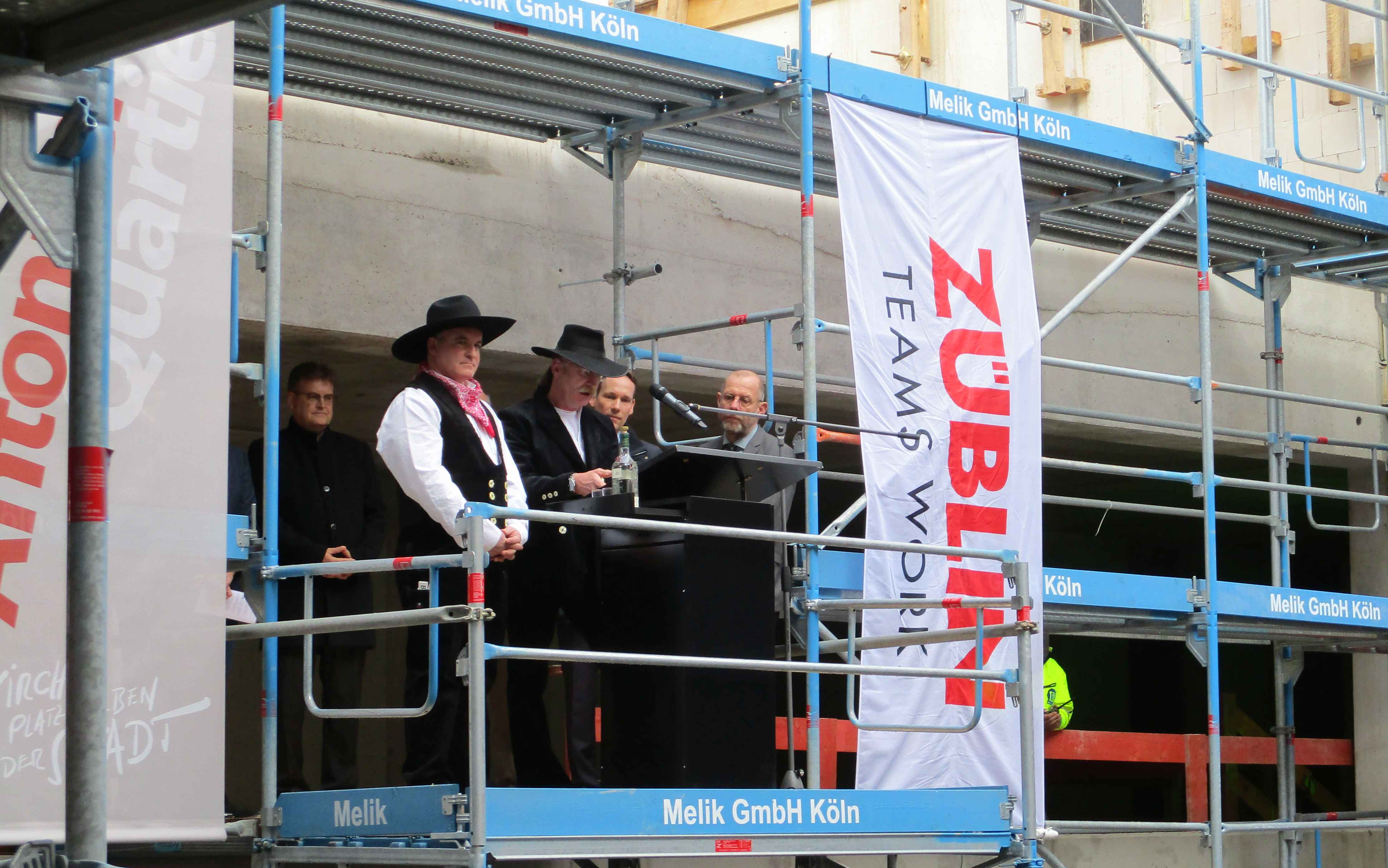 Rede zum Richtfest auf einem Gerüst auf der Baustelle im Antoniter Quartier in Köln, gehalten vom Polier. Um ihm herum sind Männer. 